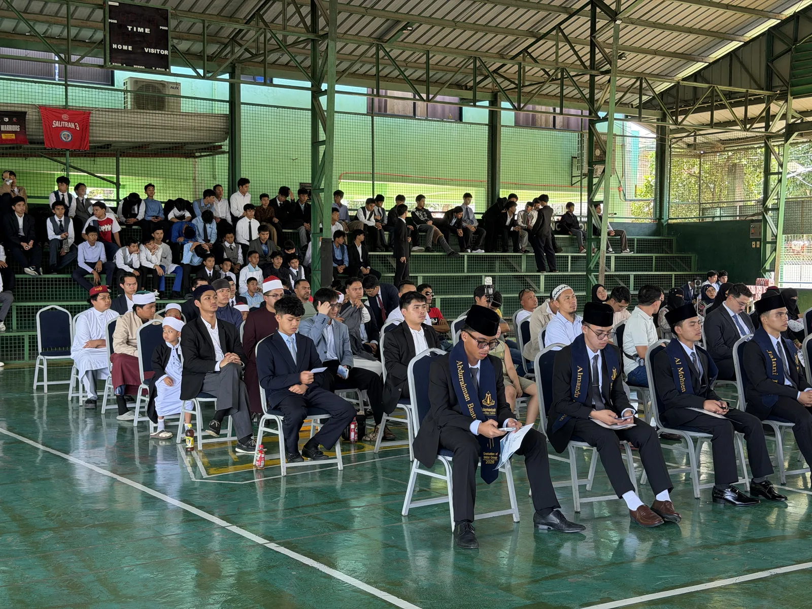 Graduates in suits seated in the front rows, with students on the bleachers behind them.