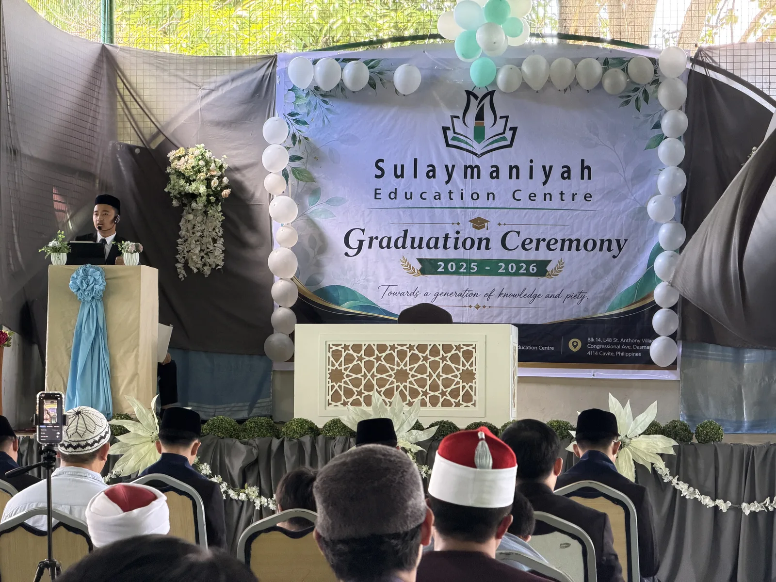 The Graduation Ceremony 2025–2026 backdrop on stage, seen over the heads of the audience.