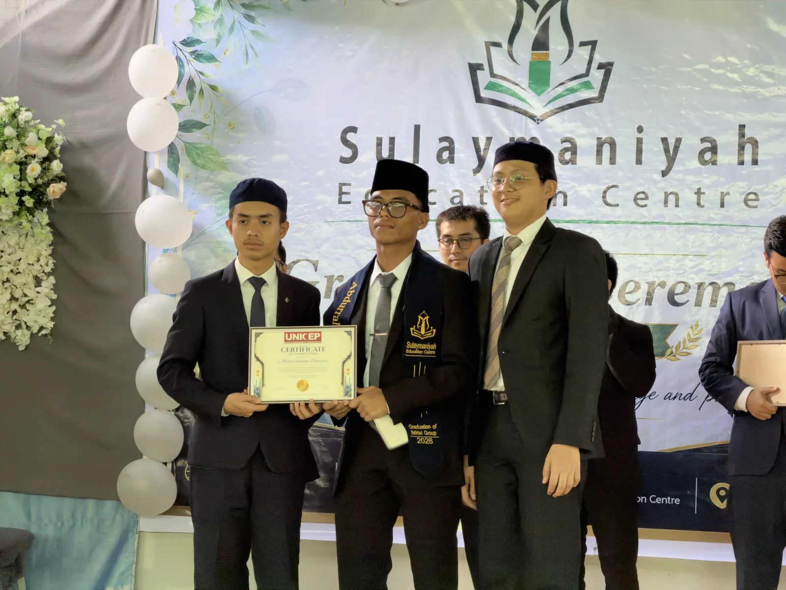 A graduate receives his certificate on stage beside two teachers, in front of the Sulaymaniyah Education Centre backdrop.