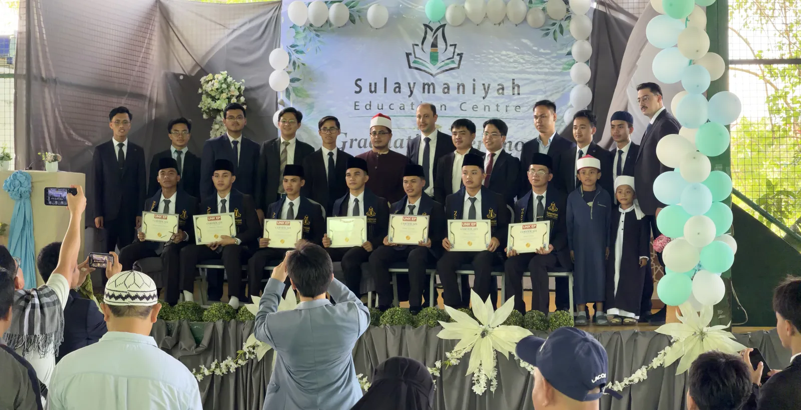 A row of graduates in suits holding their certificates on stage, with their teachers standing behind them.